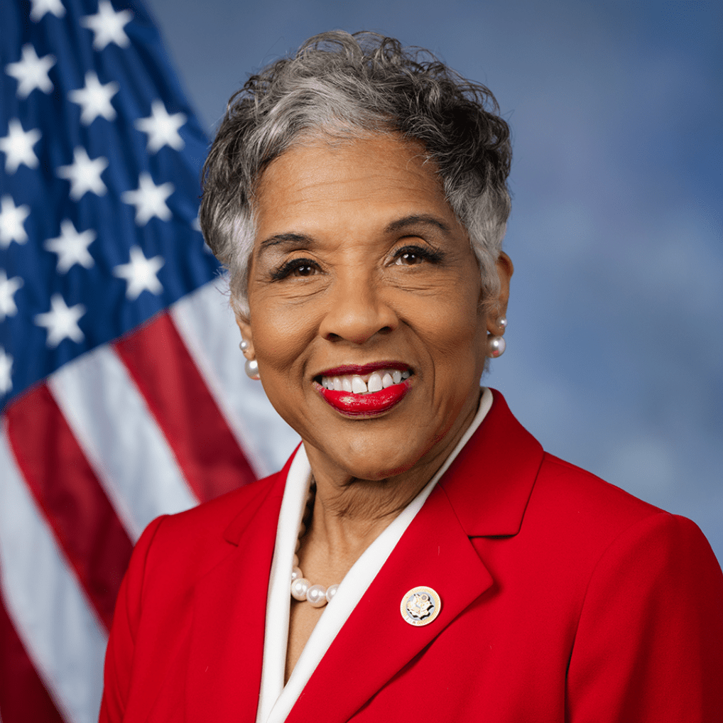A headshot of U.S. Congresswoman Joyce Beatty smiling, set against a dark blue background with the American flag visible on the left. She has short hair and is wearing a bright red blazer over a white top, accented by pearl earrings and a small lapel pin.
