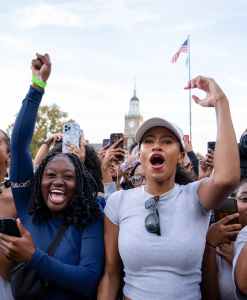 Two people cheer in a crowd of supporters, both have their hands raised, They are set against the backdrop of a Howard University building and an American flag.