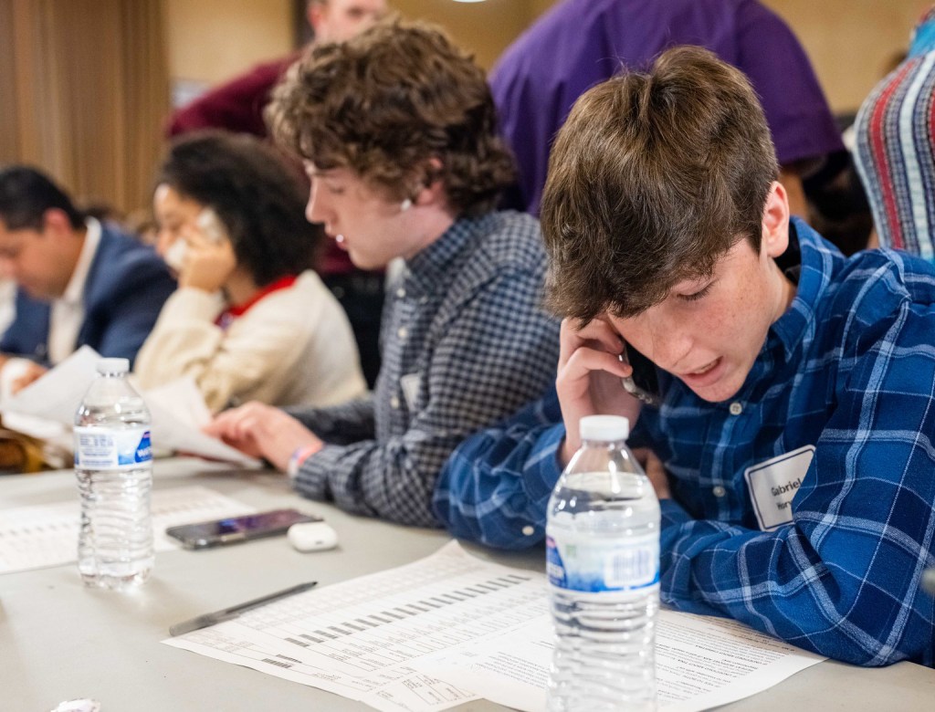 A young person wearing a blue plaid shirt and a name tag sits at a table making phone calls for the DNC, looking down at a sheet of paper with other people working in the background.