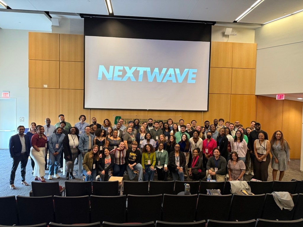 A large group of people poses and smiles together inside a university auditorium room in front of a projection screen displaying the "Nextwave" logo. The DNC's NextWave program is a free training program that serves to strengthen the next generation of campaign and party staff.