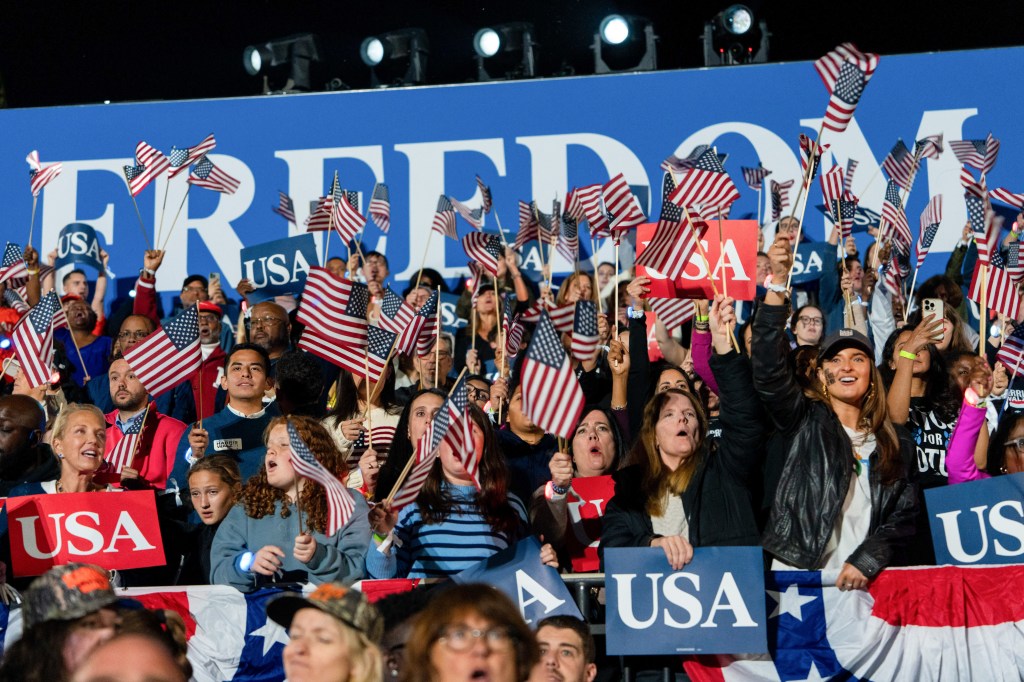 Una multitud repleta de personas vitoreando en un mitin nocturno debajo de un enorme cartel azul que dice "Freedom" en letras blancas grandes, con muchas personas sosteniendo carteles azules más pequeños que dicen "USA" y ondeando banderas estadounidenses.