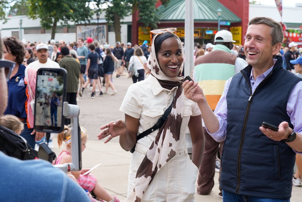 A person wearing a patterned scarf talks into a small microphone while another person in a navy vest smiles and looks on at an outdoor community event, with someone in the foreground capturing the moment on a smartphone gimbal.