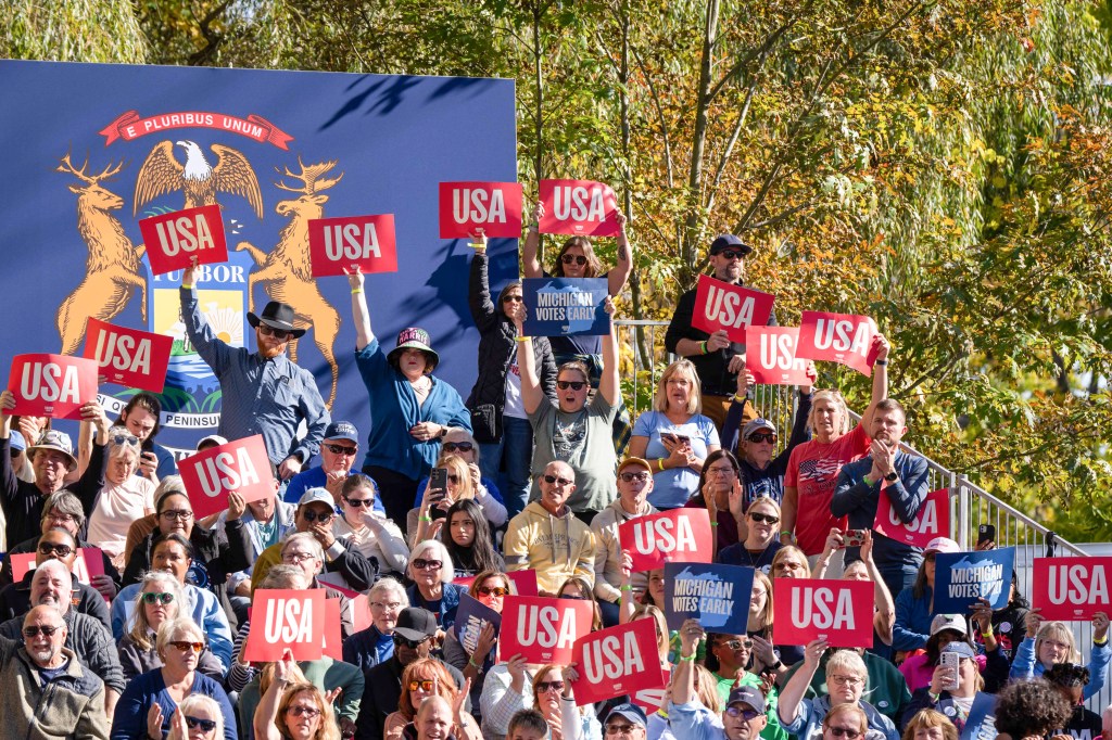 A large crowd of supporters cheers while holding up red "USA" signs at an outdoor rally in front of a blue backdrop. There are trees in the background with a mix of green and gold leaves.