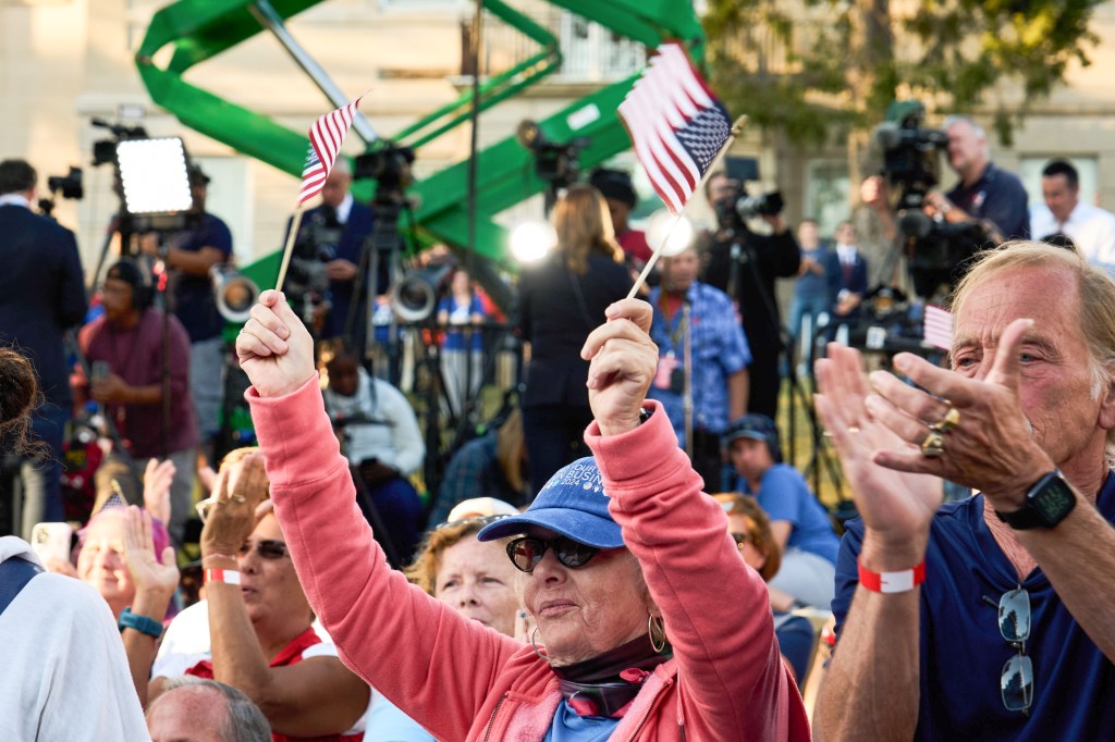 Supporters cheer at an outdoor event, with one person in a pink jacket and sunglasses holding up two small American flags, one in each hand. There is another person next to them who wears two gold rings on the left hand, and is clapping.
